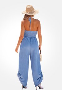 Hellblauer, strukturiertes Jumpsuit mit gerafftem Oberteil, weitem Bein und Bindebändern an den Knöcheln. Trägt einen Strohhut und eine kleine Handtasche.