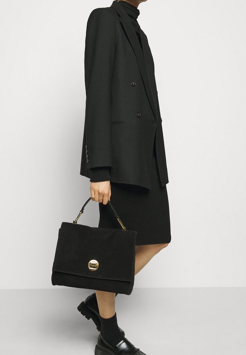 Personne portant un blazer noir, une robe noire à col roulé et des mocassins, tenant un sac à main noir avec un fermoir doré sur un fond uni.