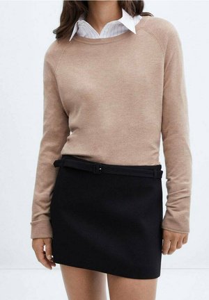 Kobieta ubrana w beżowy sweter na białą koszulę z kołnierzykiem, zestawiona z czarną mini spódnicą i cienkim czarnym paskiem.