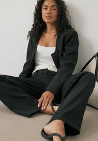 Musta räätälöity puku, jossa on istuva bleiseri ja leveälahkeiset housut, yhdistettynä valkoiseen toppiin. Housuissa on läpikuultava pinta, tasapohjaiset mustat sandaalit.