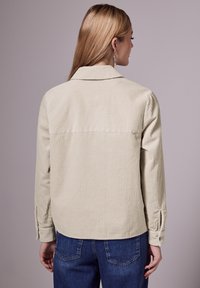 Chemise en velours côtelé beige avec une texture à côtes verticales, dotée d'un col en pointe et de poignets boutonnés, associée à un jean en denim bleu.