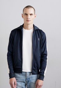 Marinblå zip-up träningsjacka med vita sidostråk, texturerad tyg, ribbade muddar och krage, bärs över en vit skjorta med ljus denimjeans.