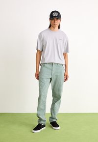 Camiseta cinza, calças listradas verdes e brancas, sapat sneakers pretos. O modelo usa um boné preto com texto branco, em pé sobre um chão verde.