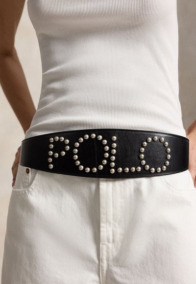 Άτομο που φοράει λευκό παντελόνι και ριμπ λευκή μπλούζα χωρίς μανίκια με μαύρη δερμάτινη ζώνη που φέρει ασημί καρφιά σχηματίζοντας τη λέξη "Polo".