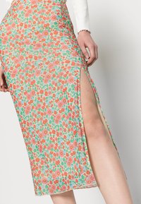 Saia floral em verde pastel, com um padrão vibrante de flores em vermelho e rosa. Fenda lateral notável e material leve e fluido.