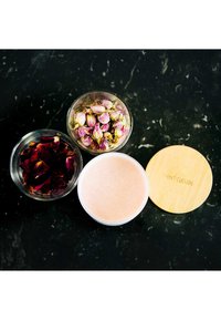 Tre contenitori di vetro trasparente contengono petali di rosa essiccati e crema rosa, con un coperchio in bambù etichettato "SAINT CLOUDS". Sfondo a macchie nere.