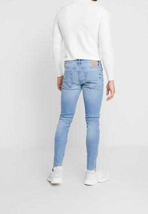 Homme portant un jean bleu clair ajusté, un pull blanc, des chaussettes blanches et des baskets blanches, debout face à un fond gris clair, dos tourné.