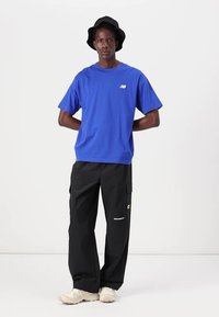 Koninklijk blauwe katoenen t-shirt met logo, gecombineerd met zwarte cargo pants met zakken en branding. Witte sneakers en een zwarte bucket hat maken de look compleet.