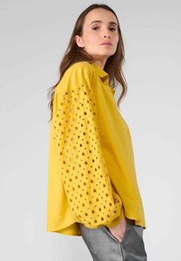 Blusa gialla con maniche a palloncino caratterizzata da motivi a cuore intagliati, realizzata in tessuto leggero, abbinata a pantaloni grigi.