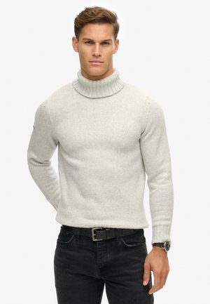 Suéter de cuello alto gris claro con un cuello y puños acanalados, hecho de un tejido de punto texturizado. Usado con jeans oscuros y un cinturón negro.