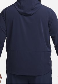 Homme portant une veste à capuche bleu foncé vue de dos, avec des manches longues et une couture au niveau du bas du dos.