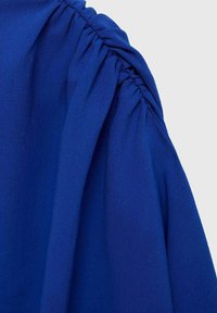 Tessuto blu elettrico con una consistenza morbida e liscia, caratterizzato da dettagli arricciati lungo la cucitura della spalla.