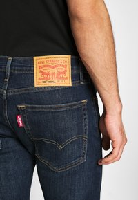 Tmavě modré džíny z denimové tkaniny mají hnědou koženou nášivku s nápisem "Levi Strauss & Co." s oranžovým prošíváním a červenou visačkou na zadní kapse.