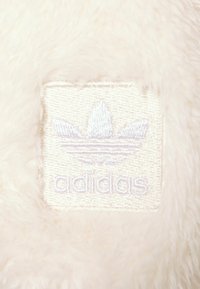 Vit fleece-lapp med broderad adidas-logotyp som har tre streck och en treklöverdesign i ljuslila. Den texturerade ytan är mjuk och fluffig.