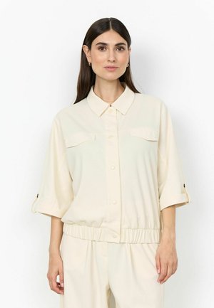 Woman with long dark hair wearing a cream buttoned shirt with rolled-up sleeves and matching pants against a plain white background.