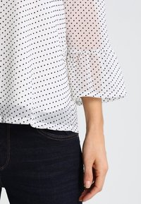 Blusa bianca con pois neri, texture trasparente, maniche a campana e orlo arricciato, abbinata a jeans in denim scuro.