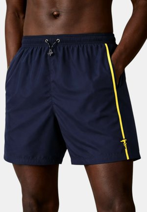 Uomo che indossa pantaloncini da bagno blu navy con strisce gialle laterali e vita regolabile con coulisse, mani in tasca.
