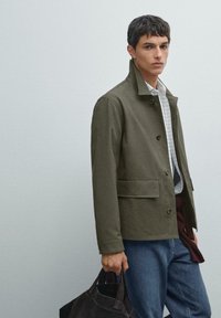 Olivenfarvet jakke med ståkrave, frontknapper og sidelommer, parret med en ternet skjorte og blå jeans. Bærer en mørk taske.