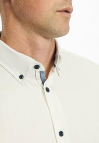 Cremfarbendes Hemd mit breitem Kragen, ausgestattet mit schwarzen Knöpfen und einem subtilen blauen und weißen gestreiften Besatz im Krageninneren.