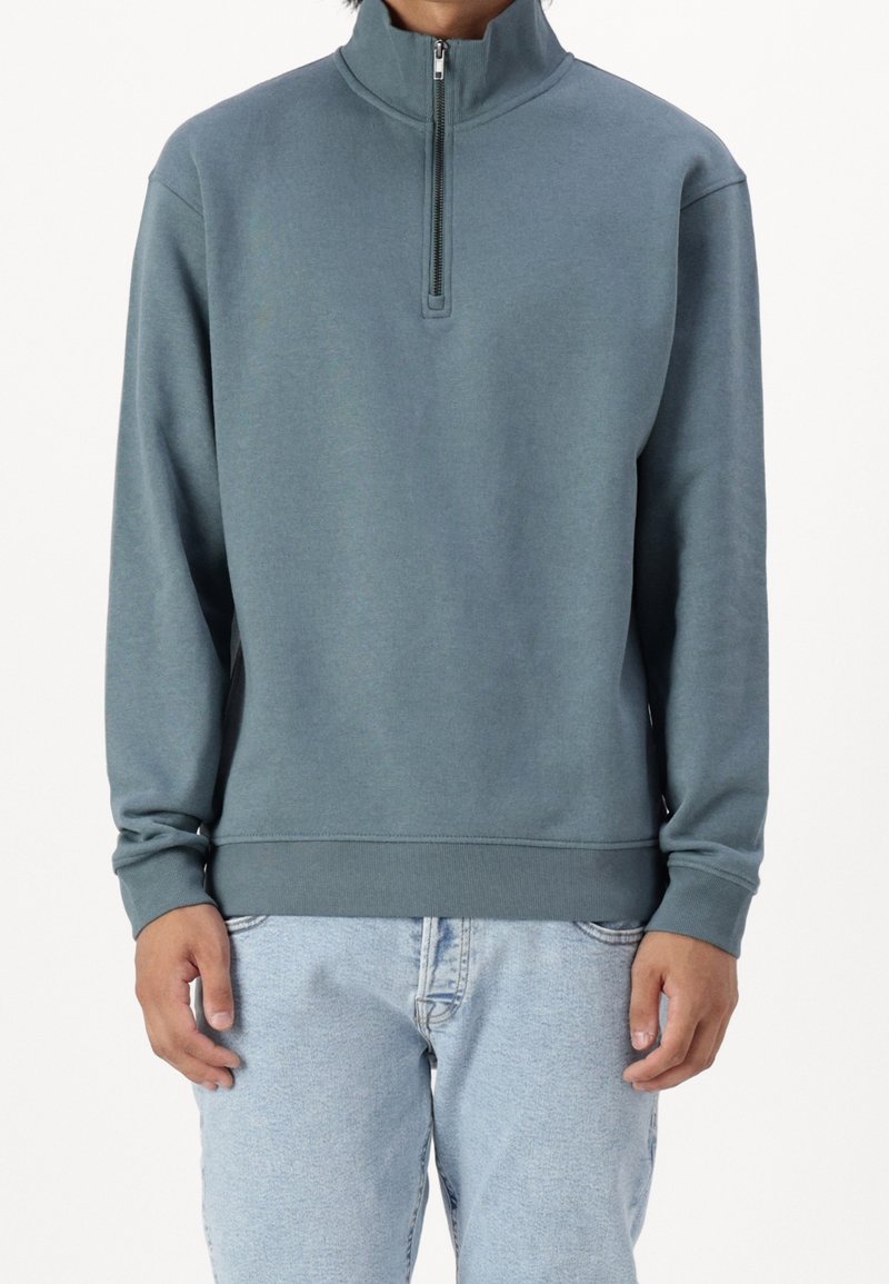 Persona vestita con una felpa blu polveroso a mezza zip e jeans azzurri, in piedi su uno sfondo bianco uniforme.