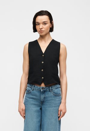 Νεαρή γυναίκα με κοντά σκούρα μαλλιά φοράει αμάνικο μαύρο γιλέκο με κουμπιά και ψηλόμεσο μπλε τζιν, στέκεται μπροστά σε απλό φόντο.