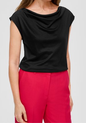 Frau trägt ein schwarzes, ärmelloses Top mit drapiertem Hals und dazu hoch taillierte, maßgeschneiderte, leuchtend pinke Hosen.