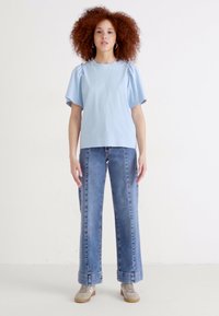 Top celeste a maniche corte con dettagli arricciati, abbinato a jeans a gamba larga in denim chiaro. La modella indossa sneaker grigie, in piedi davanti a uno sfondo neutro.