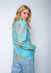 Kwiatowa koszula z długim rękawem w jasnym niebieskim kolorze z beżowymi wzorami kwiatów i liści. Posiada luźny krój i zapinane na guziki mankiety. Widoczne są dżinsowe spodnie.