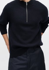 Homme portant un pull en maille côtelée noire avec une fermeture éclair argentée à moitié et un pantalon à fines rayures foncées, mains reposant derrière le dos.
