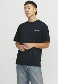 Camiseta de algodón negra con un corte holgado, escote redondo y mangas cortas. Presenta un texto de logo en blanco en la parte izquierda del pecho. Se lleva con jeans azules.