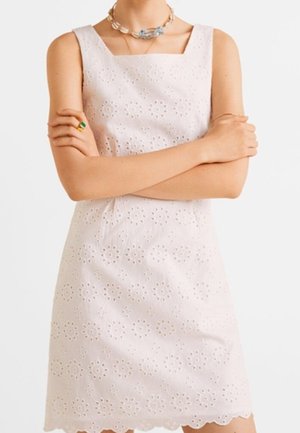 Femme portant une robe blanche sans manches avec un motif floral en broderie anglaise, un collier en forme de coquillage et une bague verte, les bras croisés sur la poitrine.