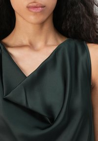 Acercamiento de una mujer que lleva una blusa sin mangas de satén verde oscuro con un escote drapeado, mostrando su cuello, hombros y labios.