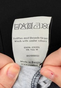 Zwart kledinglabel met textielverzorgingssymbolen, tekst in het Zweeds en het Engels, met wasinstructies en oorsprong, vastgehouden door vingers.