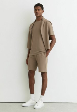 Mand står mod en hvid væg iført beige shorts og t-shirt med en matchende jakke hængende over skuldrene, hvide sokker og hvide sneakers.