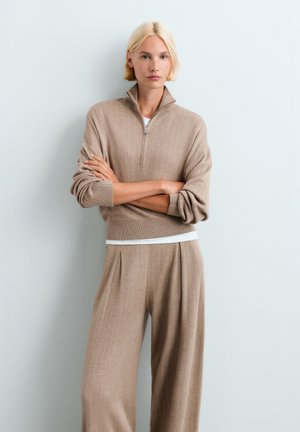 Femme aux cheveux blonds courts portant un pull zippé beige et un pantalon large assorti, debout, bras croisés, devant un mur clair uni.
