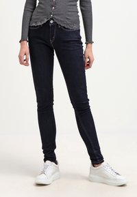 Vaqueros skinny de color azul oscuro con una textura suave, costuras en contraste y una cintura de tiro medio, combinados con zapatillas blancas para un look casual.