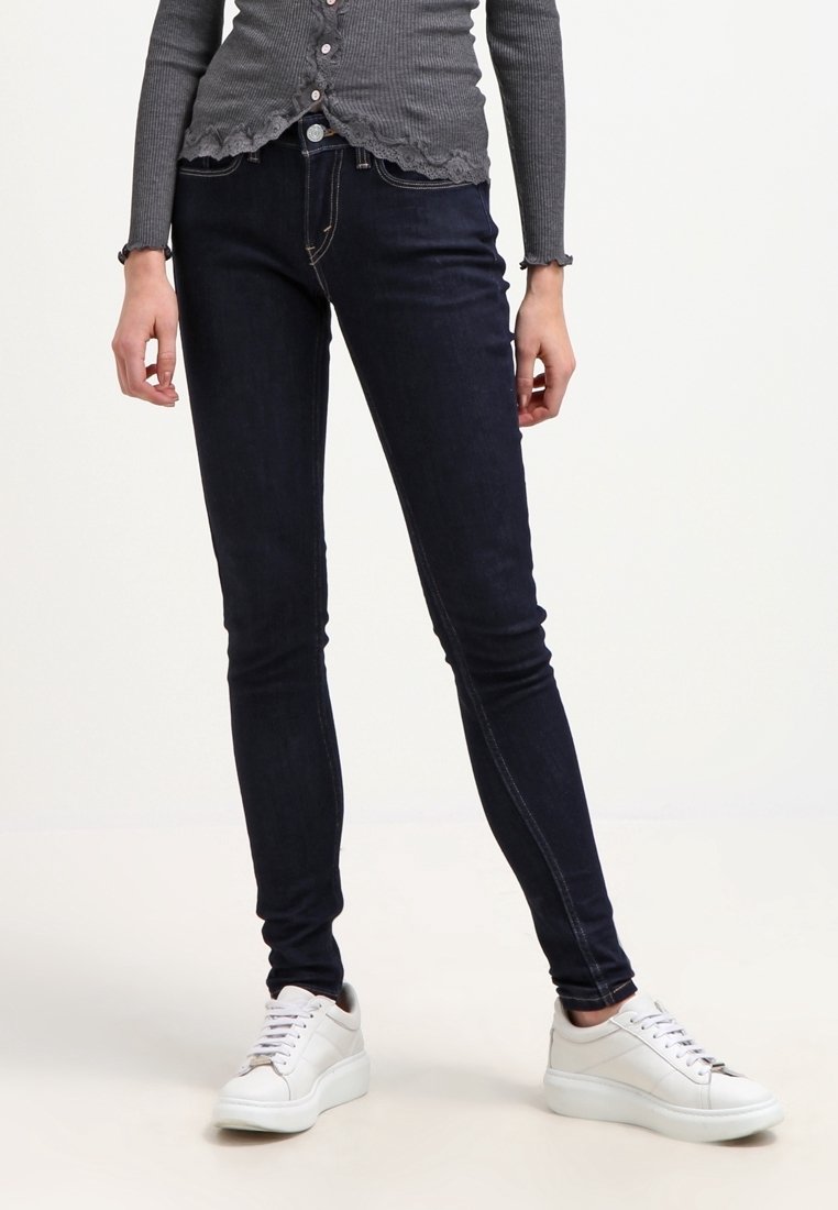 Vaqueros skinny de color azul oscuro con una textura suave, costuras en contraste y una cintura de tiro medio, combinados con zapatillas blancas para un look casual.