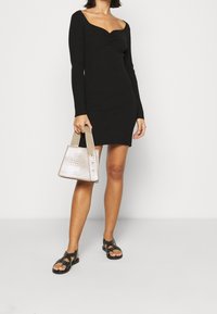 Kvinna som bär en figursydd svart långärmad miniklänning, håller en beige texturerad handväska och har på sig svarta flat sandals med nitar.