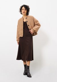 Veste courte texturée marron avec des boutons noirs, portée sur une robe cintrée marron foncé, accompagnée de bottines noires. Design simple et couleurs discrètes.