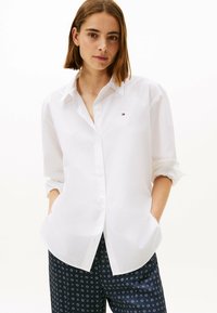 Camisa blanca de botón con cuello clásico, mangas largas arremangadas y pequeño logo bordado en el pecho. Tejido suave, corte relajado.