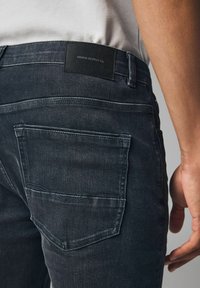 Sötét farmernadrág sima textúrával, fekete bőr címkével, rajta a "DENIM SUPPLY Co." felirattal, valamint két hátsó zsebbel, amelyek varrási részletekkel rendelkeznek.