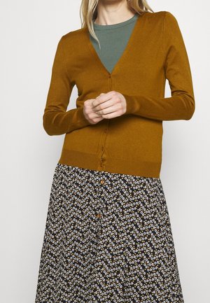 Femme portant un cardigan moutarde par-dessus un top vert et une jupe noire avec des motifs de fleurs blanches et de zigzag, debout les mains jointes.