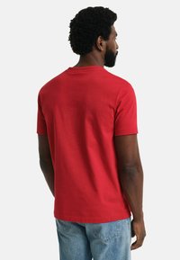 Maglietta di cotone rossa con collo a giro e maniche corte, dettagli di cucitura visibili, indossata con jeans in denim blu. Vista posteriore.