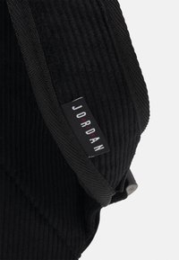 Czarna prążkowana tkanina z czarnym tkaninowym wykończeniem i czarną metką z napisem "JORDAN" w białych i czerwonych literach.