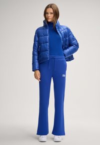 Blauer Pufferjacke mit hohem Kragen, kombiniert mit einem blauen engen Rollkragenpulli und passenden ausgestellten Jogginghosen. Weiße Sneakers runden den Look ab.
