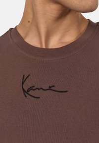 Brązowa bawełniana koszulka z okrągłym dekoltem i krótkimi rękawami, z czarnym haftowanym logiem z przodu.