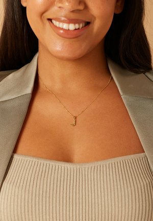 Gold necklace with a small letter "J" pendant, worn on a ribbed beige top and beneath an olive green blazer, showcasing fine chain details.