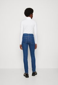 Jeans de mezclilla azul con un corte ajustado, con una cintura de altura media, bolsillos traseros y una textura suave, combinados con una blusa blanca de manga larga.