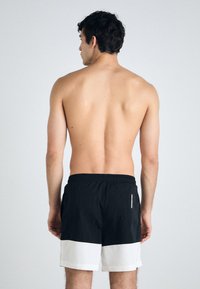 Svart svømmeshorts med en hvit horisontal stripe nederst. Lett materiale, elastisk midje og en sidelomme på baksiden.