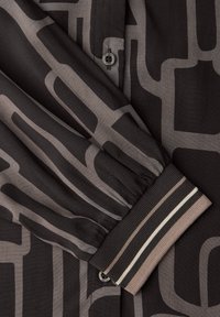 Chemise en tissu noir avec un motif abstrait gris clair, dotée d'une fermeture à boutons et de poignets élastiques ornés de détails rayés.